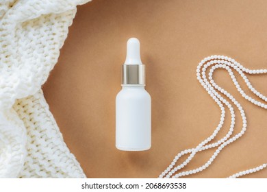 White Serum Bottle On Beige Background With Knitted Fabric And Pearl Necklace. Natural Cosmetics Concept. Top View, Flat Lay