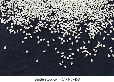 White Sesame Seeds Scattered On A Black Textured Stone Plate