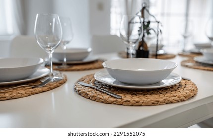 A white set dining room table with white plates, cutlery and glasses on woven place mats. There is a small bonsai in the background.