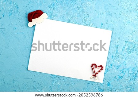 a white sheet on a blue surface in the upper corner of the sheet is fixed a paper Santa Claus hat in the lower corner sweets folded with a heart a conceptual letter to Santa Claus with New Year's wish