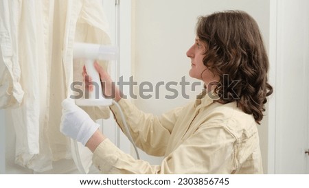 With the white shirt draped over the board, the woman uses the powerful steam to remove wrinkles from its surface. Household appliances include a trusty steamer for ironing her clothes at home.