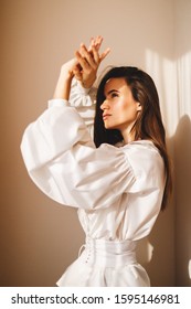 White shirt featuring long puff sleeves with cuffs and a button-up front and leather white corset in trendy minimalistic style. Light from sun on the face and shadow from hands. Woman hiding from sun.
