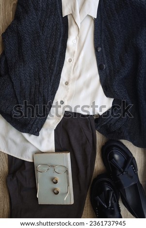 White shirt, knit cardigan, gray trousers, loafers, book, reading glasses and rings on wood background. Dark academia style outfit. Top view.