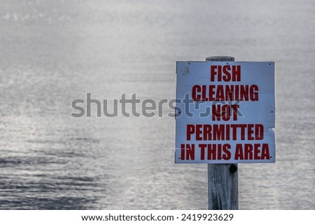 A white sign, warning notice, with red lettering indicating fish cleaning is not permitted in this area. The notice is affixed to a wooden post on a fishing wharf. The background is smooth water. 