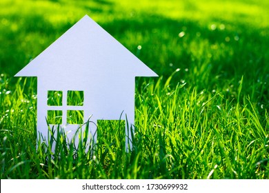 White Silhouette Of A Toy House Cut Out Of Cardboard On A Background Of Grass. A Small House Made Of White Paper. Copy Space.