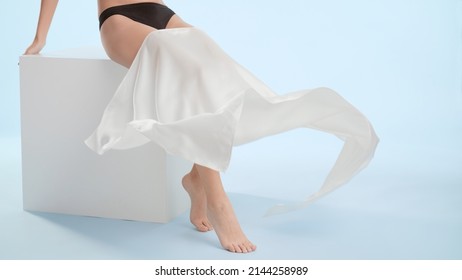 White silk cloth sliding down flawless female legs. Slim woman throws a piece of white silk cloth down her smooth legs sitting on white cube platform on blue background | Leg care concept