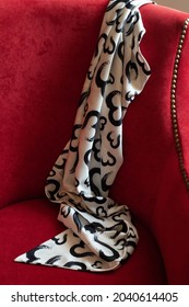 A White Silk Scarf With A Black Print Lies On A Red Chair.