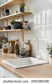 White sink in modern kitchen interior in white tones. Wooden shelves with utensils. Transparent jars with cereals on background. Contemporary apartment