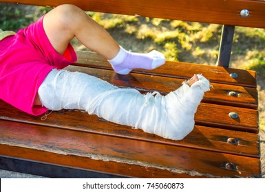 White Skin Kid With Broken Left Leg Sit On The Brown Wooden Bench Against Dry Yellow Leaves And Sunshine. Tired Child With Broken Leg In White Gypsum Rest On Park Bench.