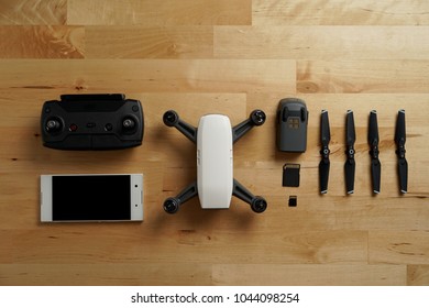 White Small Drone Is On The Wood Table. With Accessories. Spare Battery, Memory Card Remote Control, White Smartphone.