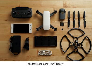 White Small Size Drone Lay Down On The Wood Table Along With Accessories Such As Battery, Battery Charger, Remote Control, Memory Card, Blades And Blade Guards