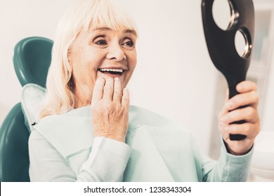 White Smile. Close Up Of Mature Smiling Woman Holding Mirror And Viewing Her New Denture While Touching Her Chin With Hand