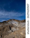 White smoke billows from the crater of the volcano, surrounded by rocky hills and green vegetation. The blue sky with wispy clouds adds a dramatic touch to the volcanic landscape.