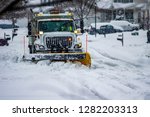 White snowplow truck with warning lights and yellow blade moving snow off of city streets while flakes are still falling