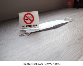 A white square plastic sign with a medical mask is placed on wooden table - black NO SMOKING sign with a picture of cigarette in red circle and Thai text saying “disobey, fine 5,000 baht”. - Shutterstock ID 2269773065