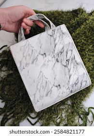 White square shape bag with gray marble pattern in hand close up. Small size minimalist woman's handbag made of PU vegan leather. Green knitted fluffy scarf and white surface at background. Accessory 