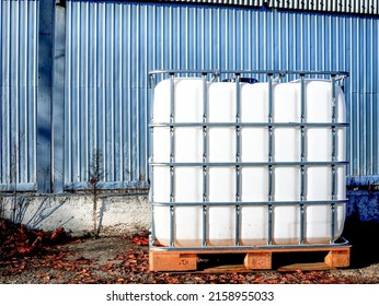 White square water tank with metal grate container for liquid standing outdoor on wooden pallet in front of a steel wall