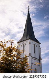 White Steeple With A Tree In Autumn Colors And A Blue Sky In A Small Village In The Country Side Of Germany, Europe