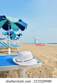 White Straw Hat On A Beach Bed With Colorful Umbrellas On A Sunny Summer Day. In The Background A Lifeboat Alongside A Lifeguard Tower On The Shore Of The Adriatic Sea, Italy.