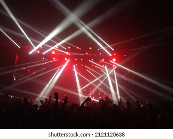 White strobe lights at a gig. A crowd of fans raising their hands at a live concert. A bunch of fans in front of strobe lights and red lights above the stage