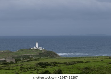 The white structure of Fanad Head Lighthouse contrasts with the deep blue of the Atlantic Ocean and green fields, highlighting the rugged coastal landscape of County Donegal, Ireland - Powered by Shutterstock