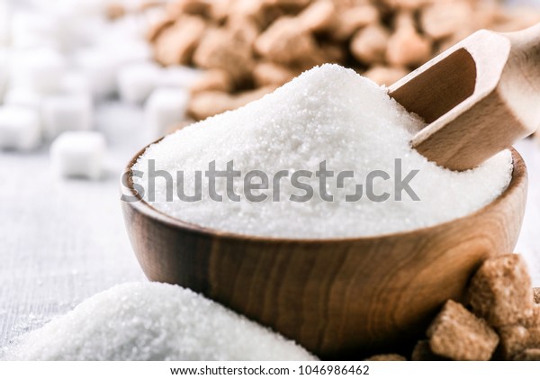 White sugar in wooden bowl\
with scoop on white table. White and brown sugar cubes in\
background.