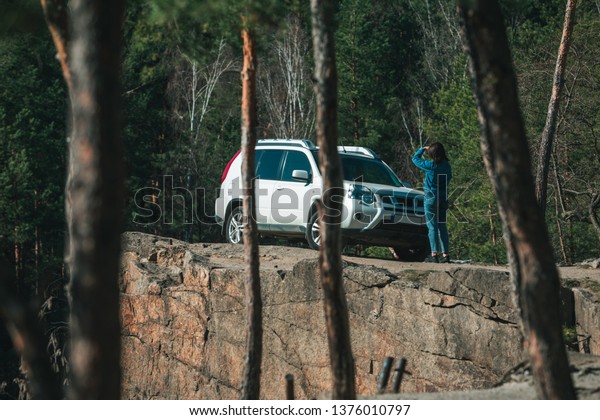 white suv car at rocky cliff young woman near it.
view through trees