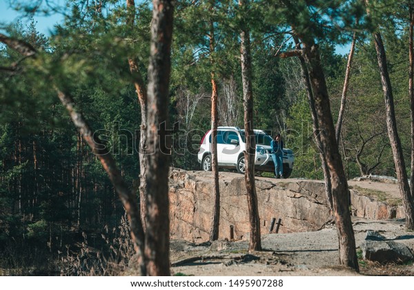 white suv car at rocky cliff young woman near it.\
view through trees