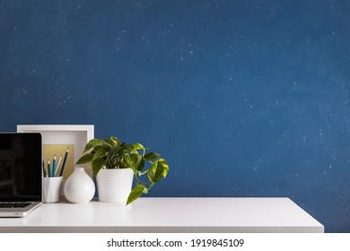 Weißer Tisch an blauer Wand mit Bürobedarf, Pflanzen, Laptop und Kaffeetasse. 