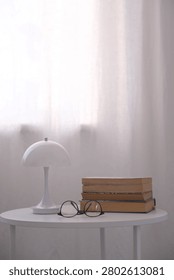 White table lamp, stack of books and eyeglasses rest on round table beside bright window light, forming quiet minimal interior scene with soft shadows, neutral tones and tidy home reading atmosphere