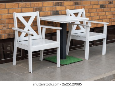 A white table and two chairs are set up on a brick sidewalk. The chairs are facing the table, and the table is empty. The scene is simple and uncluttered, with a focus on the chairs and table