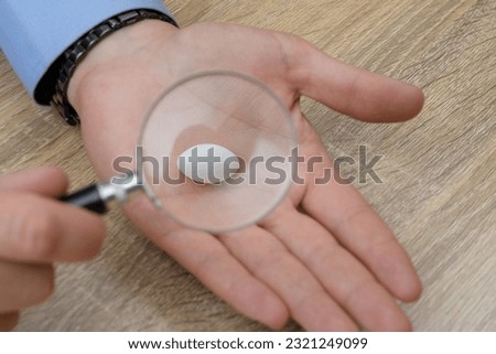 White tablet.  Medicine held in a male hand magnified with a magnifying glass