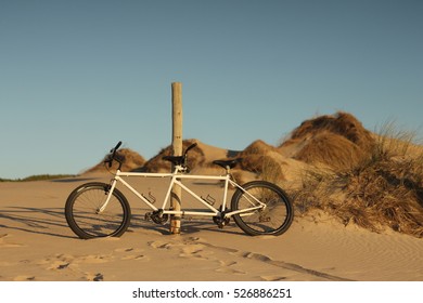 A White Tandem Bike Leaning Against A Wooden Pole On A Beach In Late Summer Afternoon