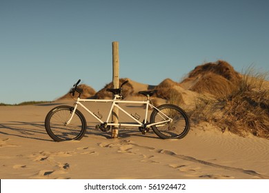 A White Tandem Bike Leaning Against A Wooden Pole On A Beach In Late Summer Afternoon. Gincho Beach, Portugal