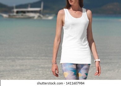 White tank top t-shirt on a young woman in leggins, front view mockup, on a beautiful beach