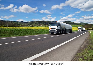 White tanker and trucks driving along the asphalt road around the yellow flowering rapeseed field in countryside. Wooded mountains in the background. Blue sky with white clouds.