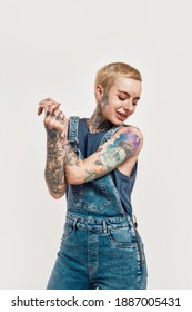 A white tattooed pierced woman holding her hands together while smiling and dancing wearing a denim overall on a white background