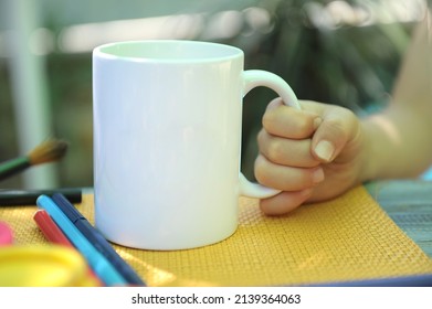 White tea mug empty space mockup, child hand with empty white cup, outdoor photo on art working desktop with colored felt pens - Powered by Shutterstock