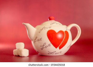 A white teapot with a red handle on the lid, black words 'love' and two red lovehearts on shown with three sugar lumps, shot against a red background with space for text, soft focus image, shallow dof
