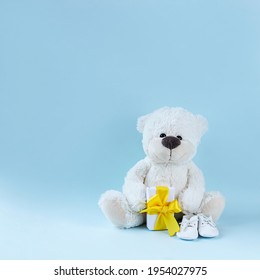 White teddy bear, baby booties and small present with yellow ribbon on light blue background. Greeting card, baby shower invitation, baby birth, gender reveal concept. Square image