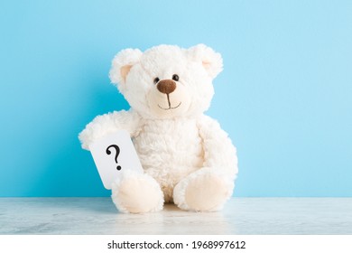 White teddy bear holding card of question sign on wooden table at light blue wall background. Pastel color. Closeup. Front view. Children issues.