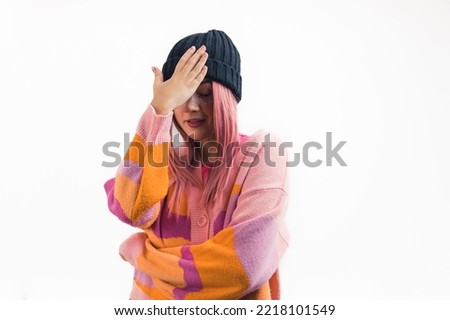 White teenage girl with pink hair wearing colorful sweater and beanie making embarassed facepalm gesture with hand to forehead. Isolated white background. Horizontal studio shot. High quality photo