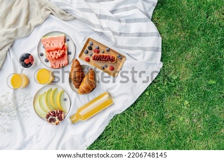 white textile table cloth, cake with berry on wooden tray, sweet croissants, juicy watermelon and ripe melon on plate. fresh juice in glassful on green grass in park at summer, top view