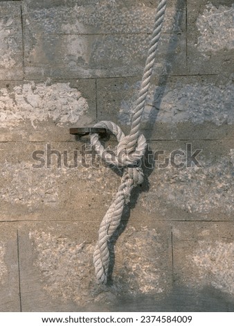 White thick rope attached to a wall rusty metal ring by a knot against a stone brick wall direct view
