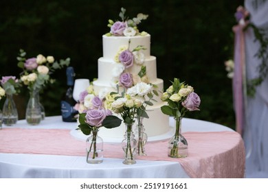 White three-tiered wedding cake decorated with roses and eustoma flowers standing on a table with a pink tablecloth outdoors - Powered by Shutterstock