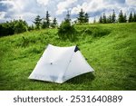 White tourist tent on sunlit, grassy hilltop, surrounded by young pine trees. Panoramic view of lush, rolling mountains under bright blue sky with scattered clouds, creating idyllic camping spot.