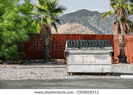 White trash dumpster in front of red wood fence with palm trees on both sides in California desert.