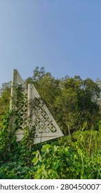 White triangular decorative structure surrounded by lush green foliage against blue sky.