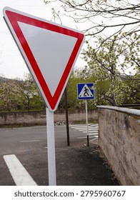 White triangular yield road sign with a red border close-up, with a pedestrian crosswalk sign and spring blossoms blurred in the background.