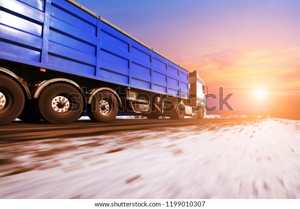 White truck and a blue trailer with space\
for text driving fast on the winter countryside road with snow\
against blue sky with clouds and bright\
sunset